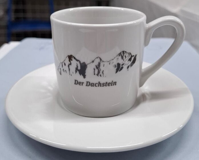 Weiße Porzellan-Espressotasse mit schwarzem Dachstein-Motiv und passender Untertasse auf hellem Tisch | © Planai-Hochwurzen-Bahnen Gesellschaft m.b.H.
