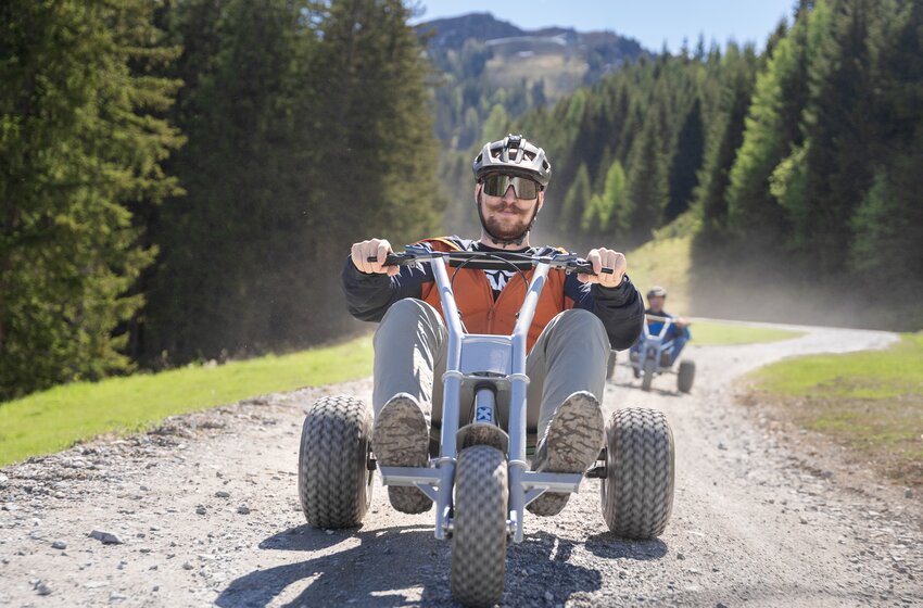 Ein Mann fährt mit einem Mountaincart eine kurvige Schotterstraße hinunter, umgeben von Wald und Wiese bei Sonnenschein | © Dorfgasteiner Bergbahnen AG