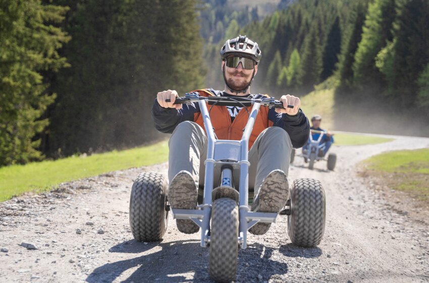 Ein Mann fährt mit einem Mountaincart eine kurvige Schotterstraße hinunter, umgeben von Wald und Wiese bei Sonnenschein | © Dorfgasteiner Bergbahnen AG