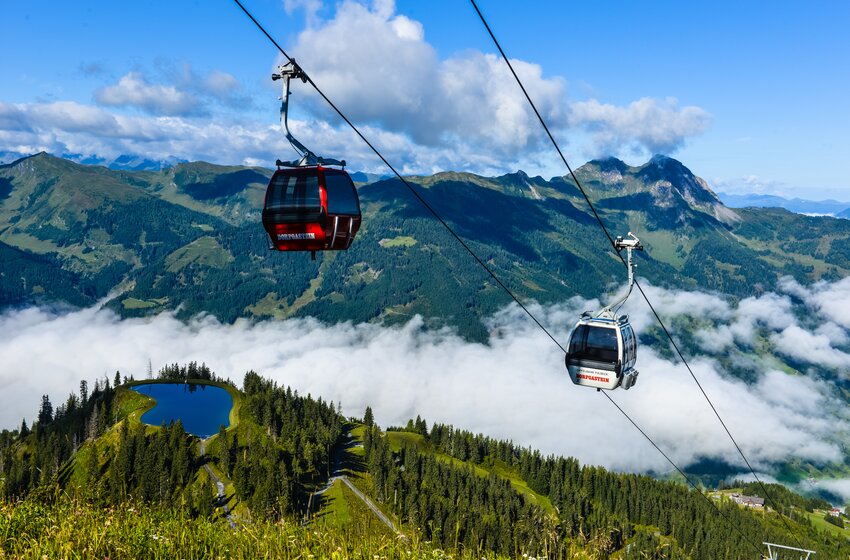 Zwei Gondeln fahren über Dorfgastein, darunter Wälder, ein Teich und Wolken, im Hintergrund Alpenpanorama. | © Dorfgasteiner Bergbahnen AG