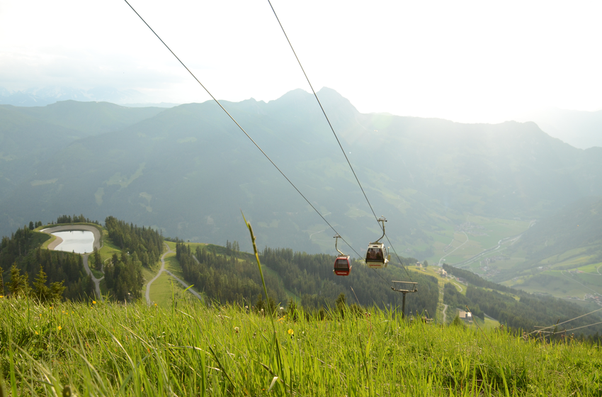 Two gondolas glide above green alpine meadows with a reservoir and wide mountain panorama in sunny conditions. | © Dorfgasteiner Bergbahnen AG