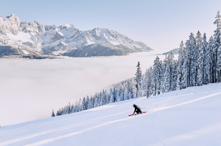 Ein Skifahrer fährt eine präparierte Piste hinab, dahinter verschneite Berge und Nebelmeer. | © Bergbahnen Filzmoos GmbH