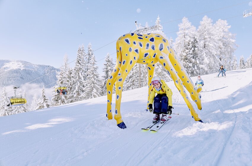 Ein Kind fährt lachend Ski durch eine gelbe Giraffenfigur, dahinter verschneite Landschaft. | © Bergbahnen Filzmoos GmbH