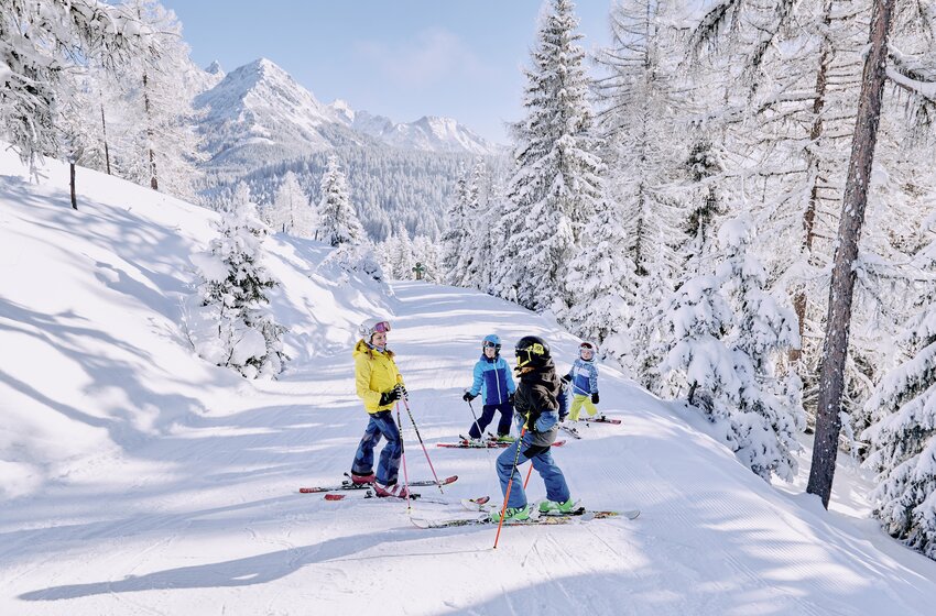 Vier Kinder stehen auf einer verschneiten Waldpiste, umgeben von Bäumen und Bergen im Winter. | © Bergbahnen Filzmoos GmbH