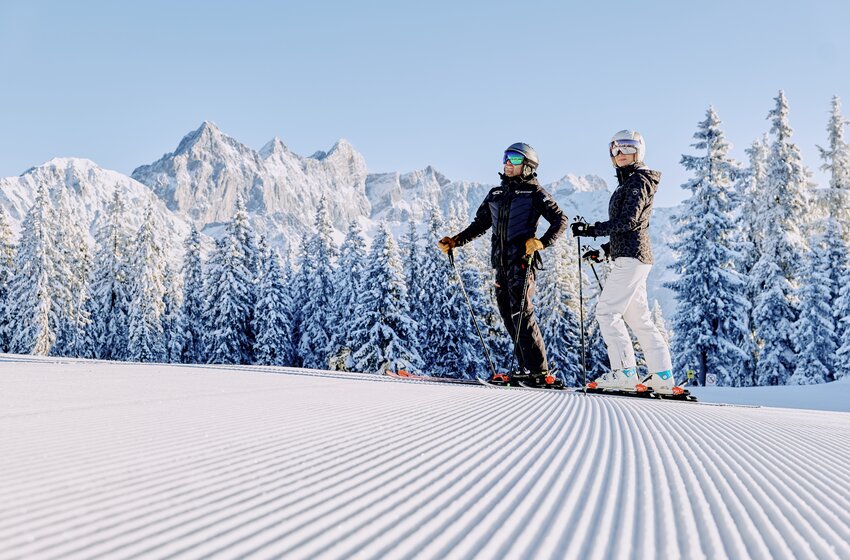 Zwei Skifahrer stehen auf gerillter Piste vor verschneiten Bäumen und dem Dachsteinmassiv im Hintergrund. | © Bergbahnen Filzmoos GmbH