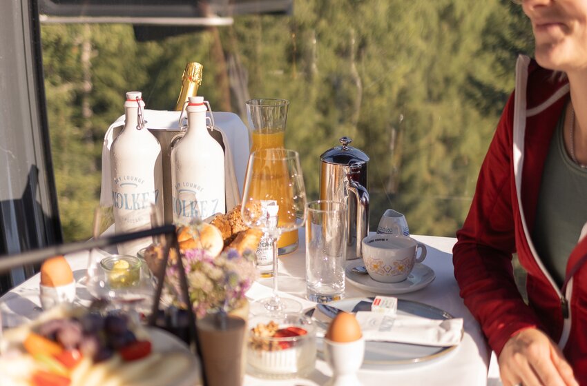 Frau sitzt an gedecktem Tisch mit Frühstück und Orangensaft in einer Berggondel vor grünem Waldhang | © Großarler Bergbahnen GmbH & Co KG