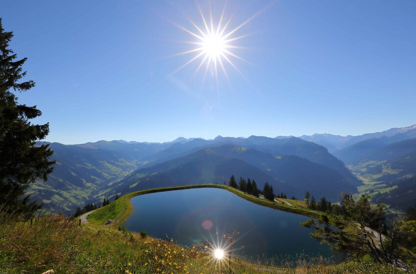 Bergsee mit Spiegelung der Sonne oberhalb des Großarltals, mit Blick auf grüne Hügel und entfernte Gipfel | © Grossarler Bergbahnen GmbH & Co KG