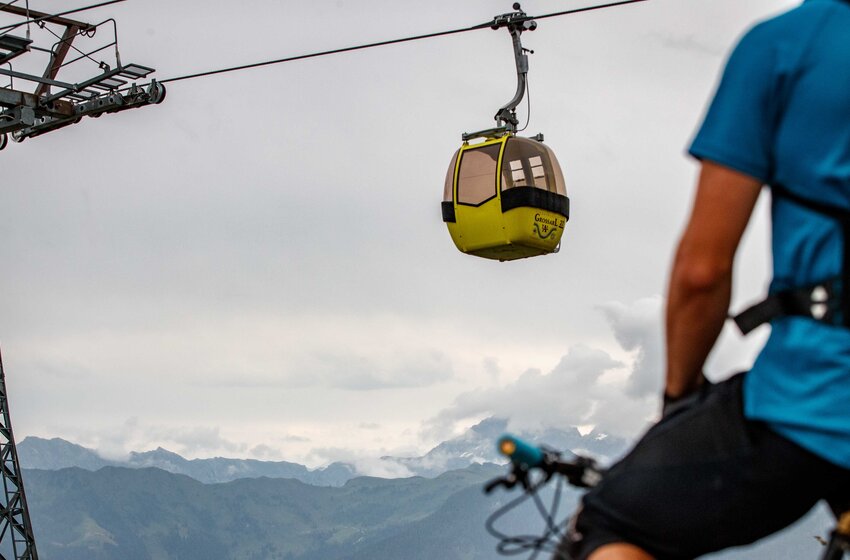 Radfahrer mit Helm blickt auf gelbe Gondel der Großarlbahn vor bewölktem Himmel und alpinem Bergpanorama | © Grossarler Bergbahnen GmbH & Co KG