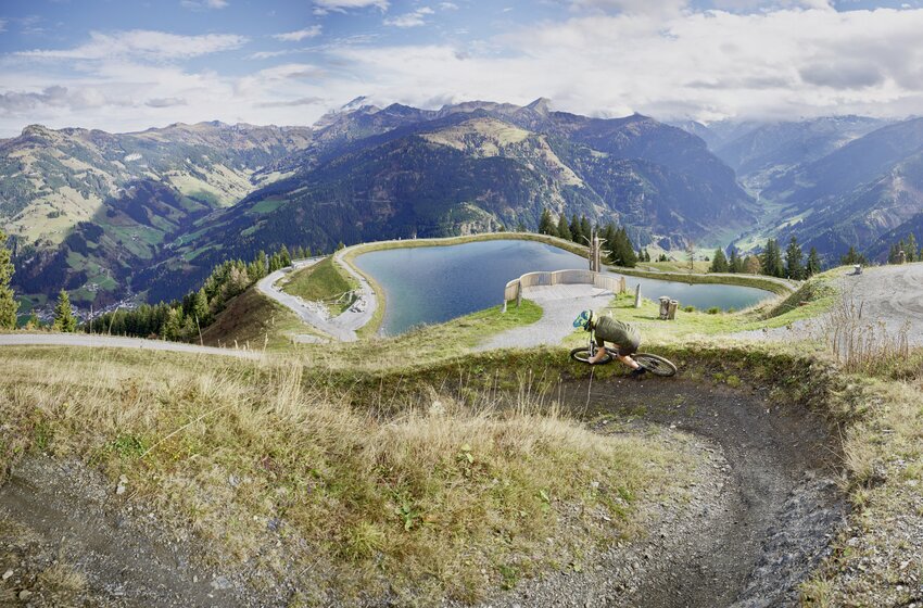 Mountainbiker fährt durch Kurve oberhalb eines Speicherteichs mit Alpenpanorama unter locker bewölktem Himmel. | © Großarler Bergbahnen GmbH & Co KG