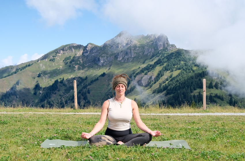 A woman meditates cross-legged on a yoga mat on a meadow in Grossarl Valley with green mountains behind her. | © Grossarler Bergbahnen GmbH & Co KG