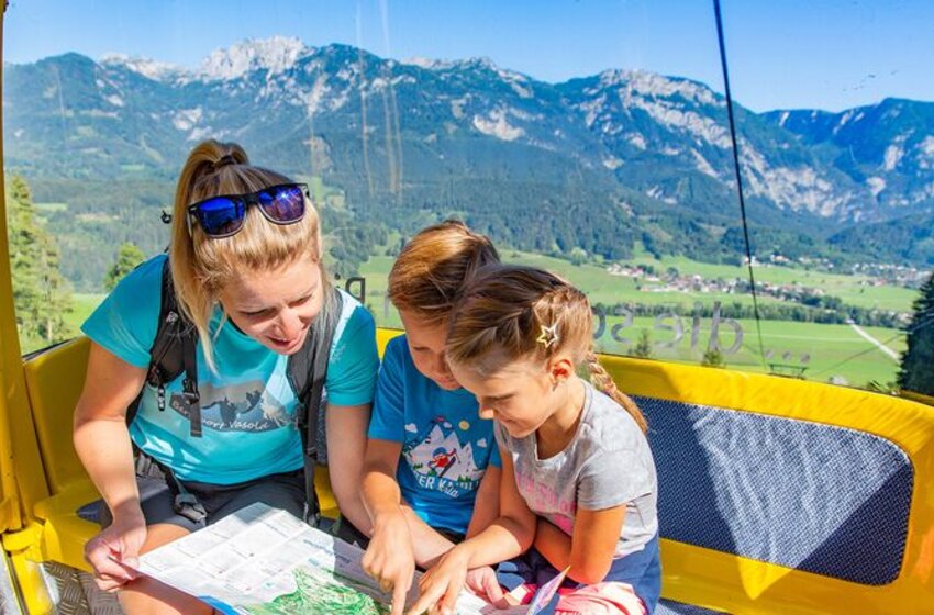 Eine Mutter und zwei Kinder betrachten eine Wanderkarte in einer gelben Gondel mit Blick auf grüne Täler und Berglandschaft im Sommer. | © Hauser Kaibling Seilbahn- und Liftges.m.b.H. & Co KG