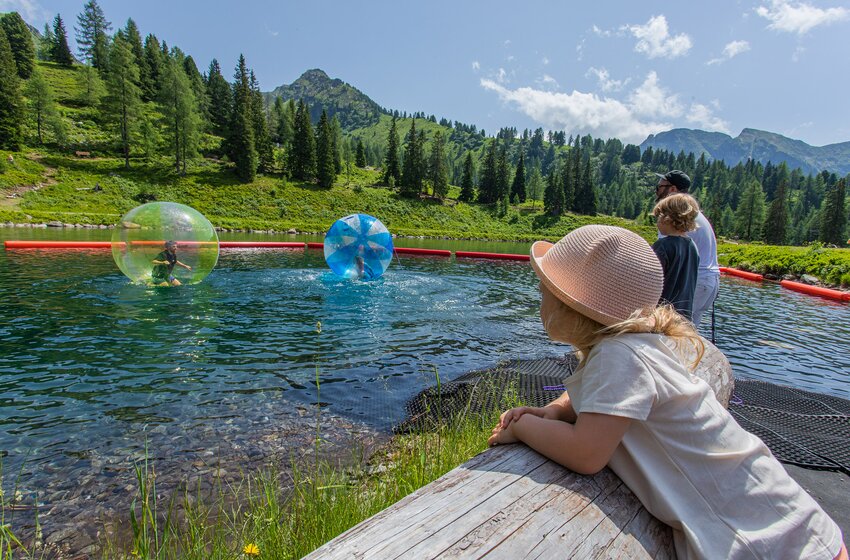 Kinder laufen in Wasserbällen über Bergsee, während Familie am Ufer in sonniger Alpenlandschaft zusieht | © Hauser Kaibling Seilbahn- und Liftges.m.b.H. & Co KG