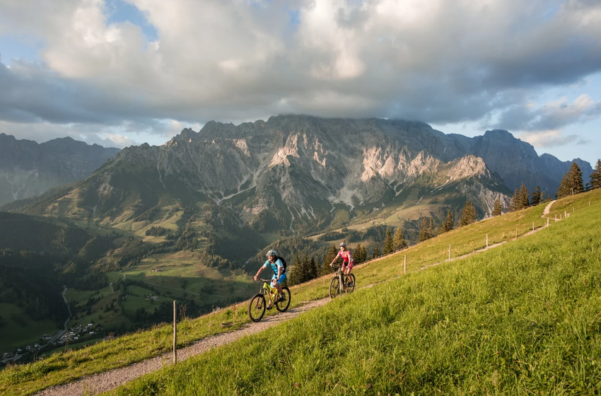 Zwei Mountainbiker fahren auf schmalem Trail über Almwiesen am Hochkönig mit Blick auf markante Berggipfel und Abendwolken | © Hochkönig Bergbahnen GmbH