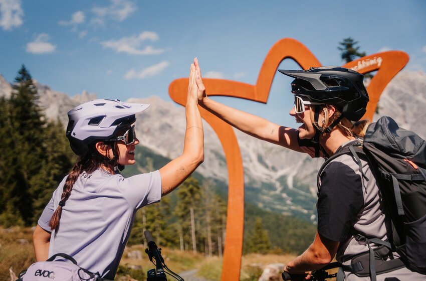Zwei Mountainbikerinnen geben sich vor Alpenkulisse mit Helmen und Fahrrädern ein High Five | © Hochkönig Bergbahnen GmbH