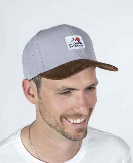 Mann mit grauem Baseballcap mit Logo und braunem Schirm trägt weißes T-Shirt vor hellem Studiohintergrund | © Ski amadé GmbH / Lorenz Masser