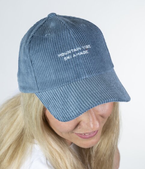 Blonde Frau trägt blaues Cord-Baseballcap mit Stickerei und blickt nach unten vor hellem Studiohintergrund | © Ski amadé GmbH / Lorenz Masser