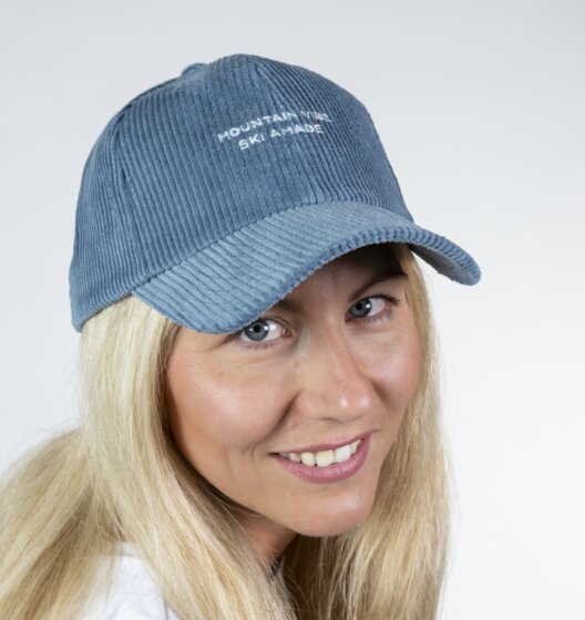 Blonde Frau mit blauem Cord-Baseballcap und weißem Shirt blickt lächelnd zur Seite vor hellem Studiohintergrund | © Ski amadé GmbH / Lorenz Masser