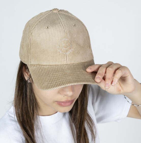 Mädchen mit beigem Cord-Baseballcap und weißem T-Shirt hält den Schirm der Kappe vor hellem Studiohintergrund | © Ski amadé GmbH / Lorenz Masser