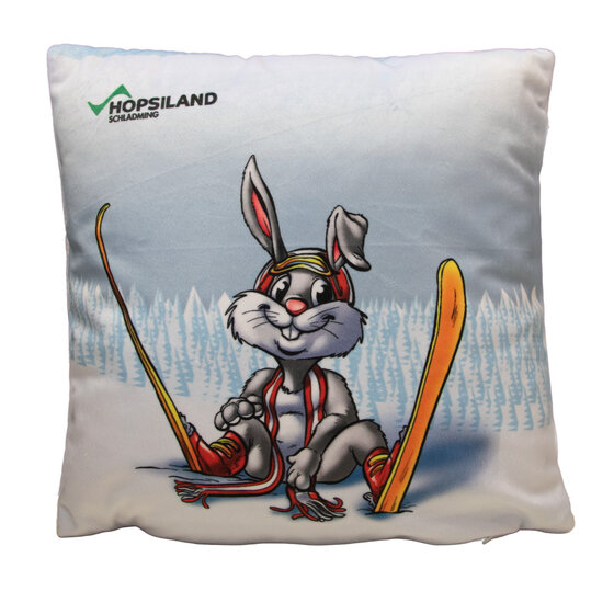 Quadratisches Kissen mit Hase Hopsi in Skiausrüstung vor verschneitem Wald, gelbe Ski und Aufdruck 'Hopsiland' | © Planai-Hochwurzen-Bahnen GmbH