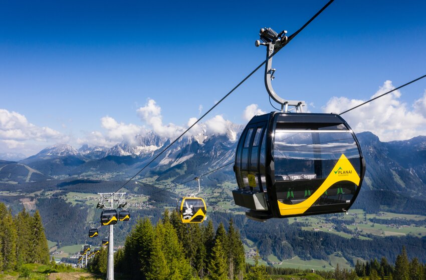 Moderne Planai-Gondeln schweben über grüne Berghänge mit Blick auf das Dachsteinmassiv und sonnige Täler | © Planai-Hochwurzen-Bahnen GesmbH