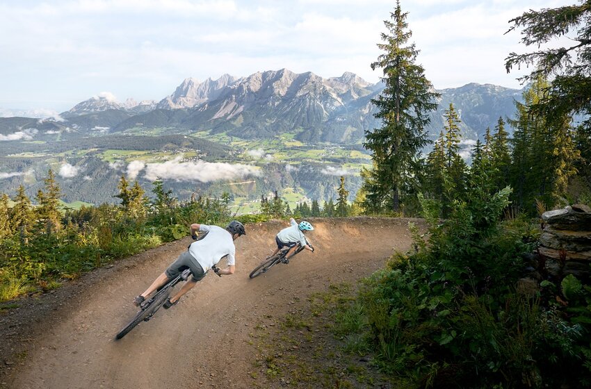 Zwei Mountainbiker auf einem kurvigen Trail mit weitem Blick auf grüne Täler, Wälder und das Dachsteinmassiv | © Planai-Hochwurzen-Bahnen GesmbH