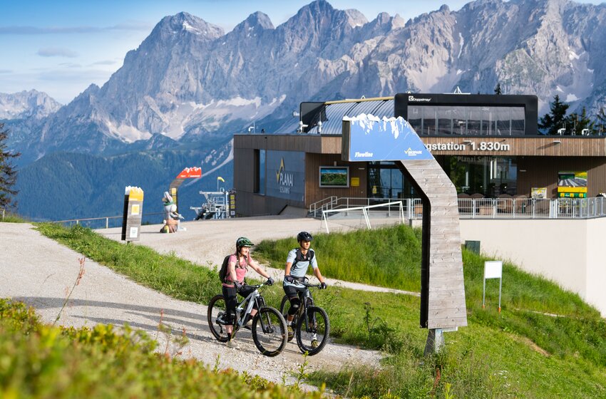 Zwei Mountainbiker stehen am Flowline-Start vor der Planai-Bergstation auf 1830 m, mit dem Dachsteinmassiv im Hintergrund | © Josh Absenger