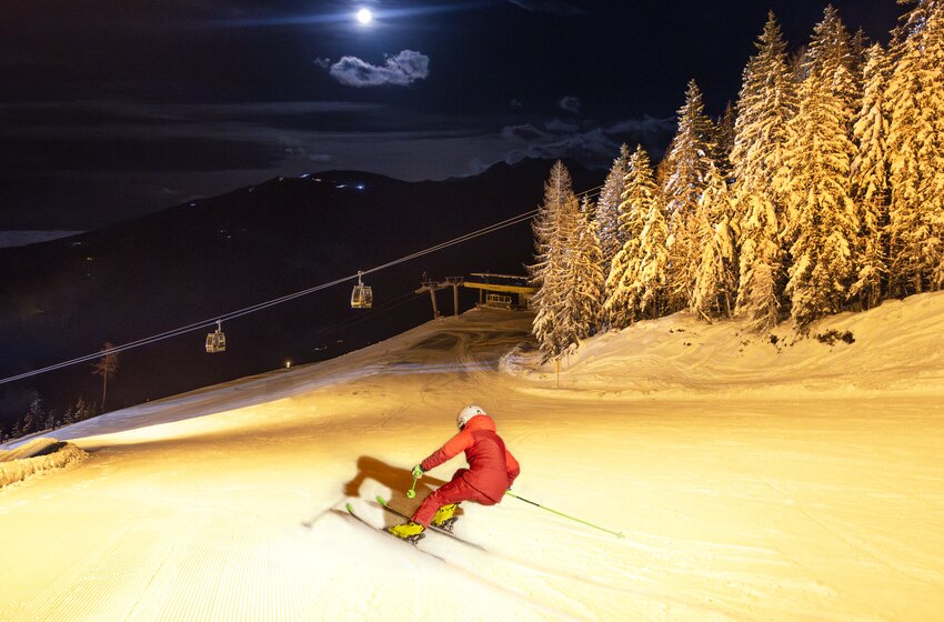Skifahrer in rotem Anzug fährt nachts auf beleuchteter Piste, umgeben von verschneiten Bäumen und Gondelbahn bei Vollmond. | © Martin Huber