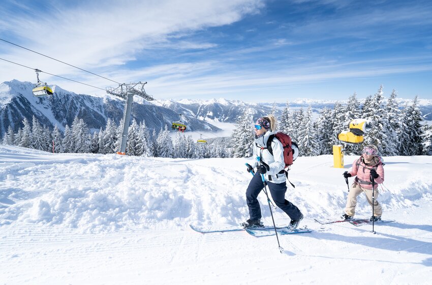 Zwei Skitourengeherinnen steigen bei Sonnenschein über eine verschneite Planai-Piste mit Blick auf die Berge.