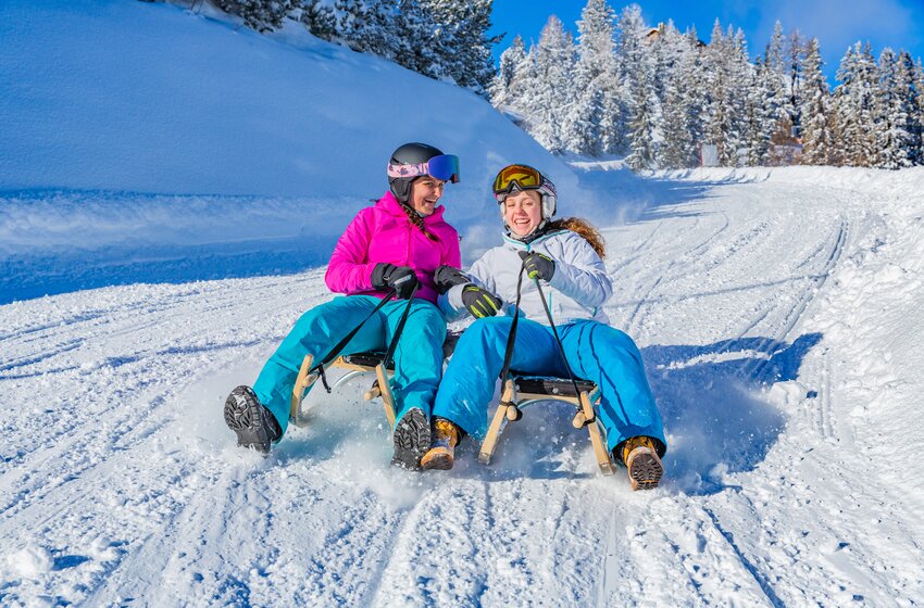 Zwei lachende Frauen rodeln nebeneinander eine verschneite Winterpiste hinunter, Schnee spritzt auf. | © René Eduard Perhab