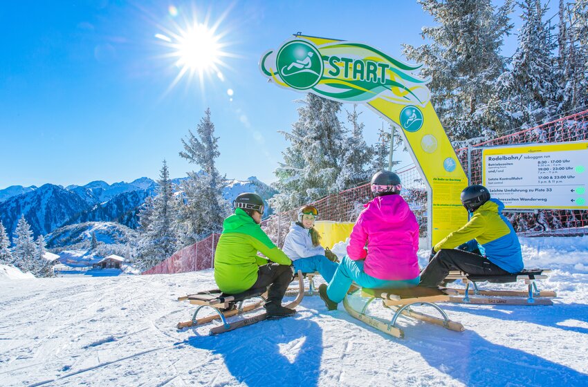 Vier Rodler:innen sitzen an sonniger Rodelbahn-Startlinie mit Blick auf verschneite Berge. | © René Eduard Perhab