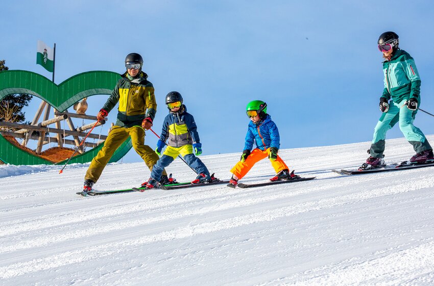 Familie fährt gemeinsam im Pflug eine sonnige Piste hinab, im Hintergrund grünes Holzherz mit Spielplatz am Rittisberg. | © Rittis-Lift Engelhardt GmbH & Co KG