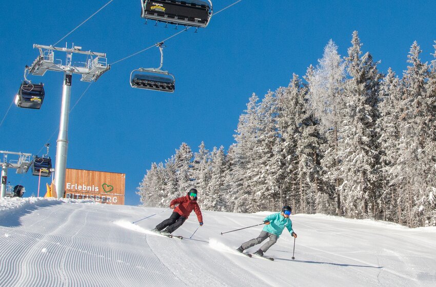 Zwei Skifahrer fahren bei Sonne auf der Piste unter der Erlebnisbahn Rittisberg, daneben verschneiter Wald. | © Rittis-Lift Engelhardt GmbH & Co KG