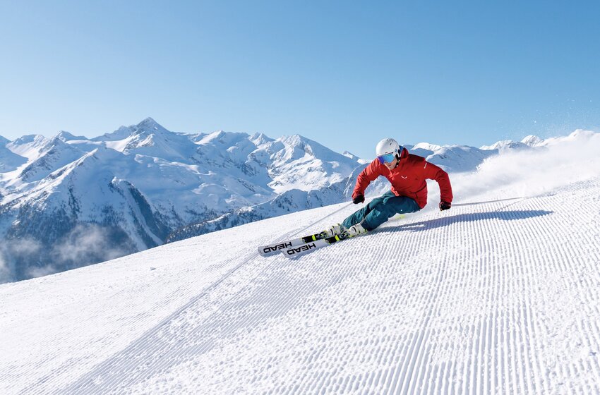 Skifahrer in roter Jacke carvt über frisch präparierte Piste vor verschneiten Gipfeln bei blauem Himmel. | © Gasteiner Bergbahnen AG