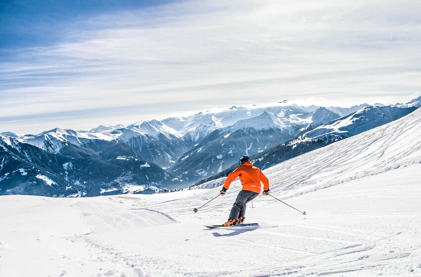 Skifahrer in oranger Jacke fährt auf breiter Piste mit Ausblick auf verschneite Täler und hohe Alpengipfel. | © Gasteiner Bergbahnen AG