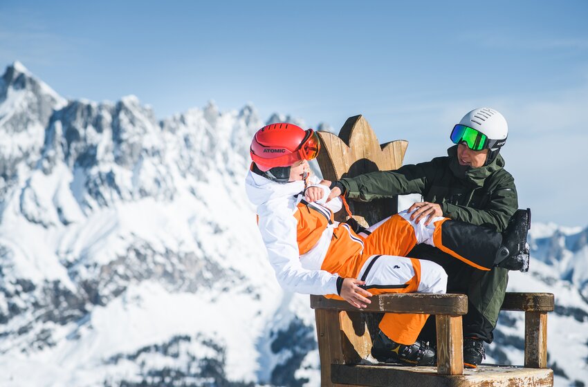 Zwei Skifahrer scherzen auf einer Holzbank mit Bergpanorama im Hintergrund am Hochkönig.  | © Hochkönig Bergbahnen GmbH