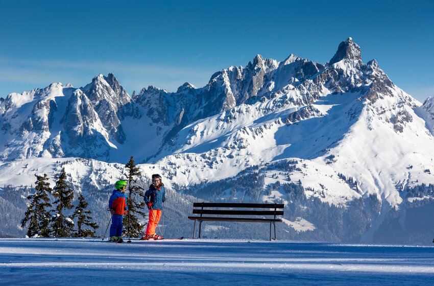 Zwei Kinder auf Skiern vor einer Bank mit spektakulärem Panorama verschneiter Alpengipfel. | © Salzburger Sportwelt Amadé GmbH