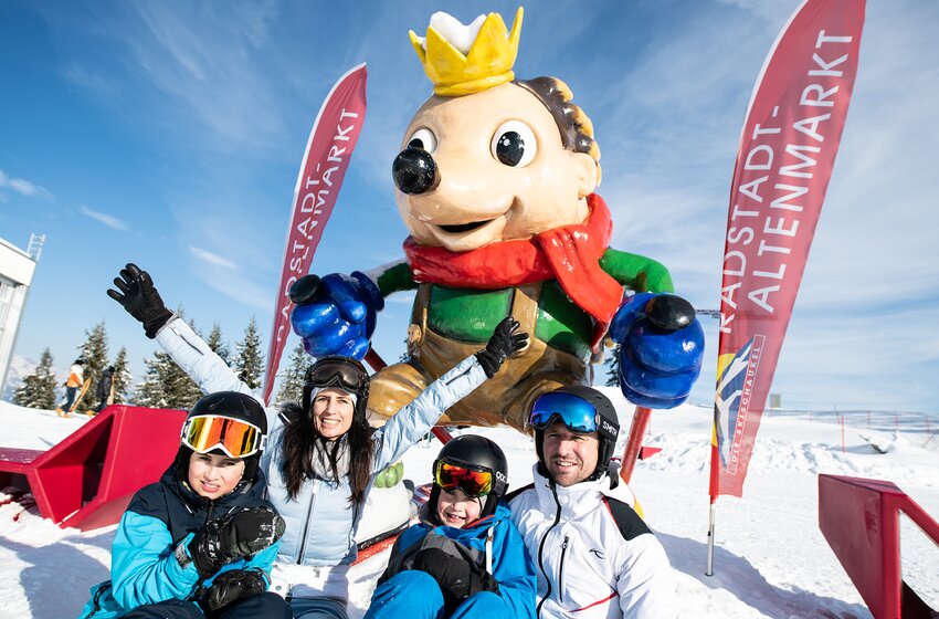 Familie in Skikleidung posiert fröhlich vor dem Maskottchen Kinigl im Skigebiet Radstadt-Altenmarkt. | © Zauchensee Liftgesellschaft Benedikt Scheffer GmbH