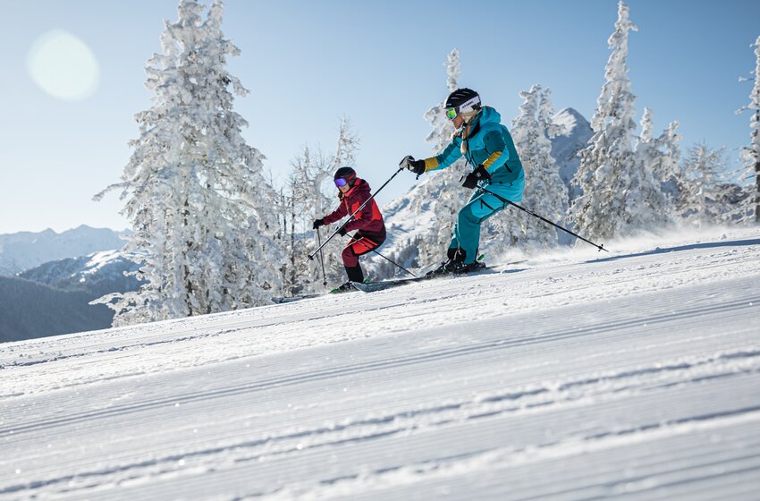 Zwei Skifahrerinnen fahren nebeneinander auf einer frisch präparierten Piste umgeben von verschneiten Bäumen und Bergen. | © Reiteralm Bergbahnen GmbH & Co.KG