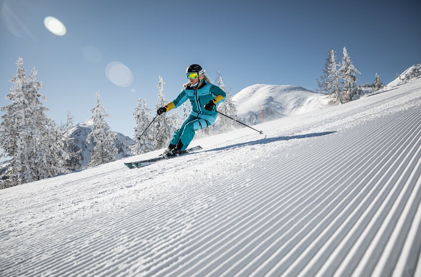 Skifahrerin in türkisfarbener Kleidung fährt auf frisch präparierter Piste bei blauem Himmel und Sonnenschein. | © Reiteralm Bergbahnen GmbH & Co.KG