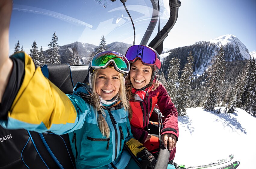 Zwei lächelnde Skifahrerinnen machen ein Selfie im Sessellift vor verschneiten Bäumen und sonniger Bergkulisse. | © Reiteralm Bergbahnen GmbH & Co.KG