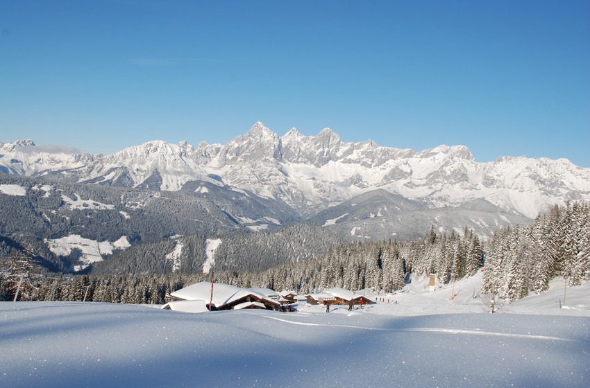 Verschneite Hütten und Wälder mit imposantem Alpenpanorama unter blauem Himmel. | © Reiteralm Bergbahnen GmbH & Co.KG