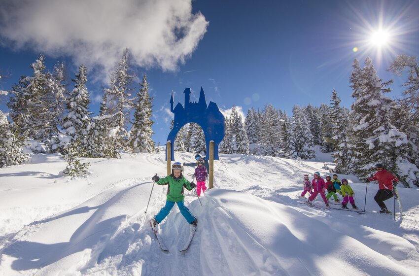 Kinder fahren Ski durch ein blaues Märchentor im Schnee bei Sonnenschein und tiefblauem Himmel. | © Reiteralm Bergbahnen GmbH & Co.KG