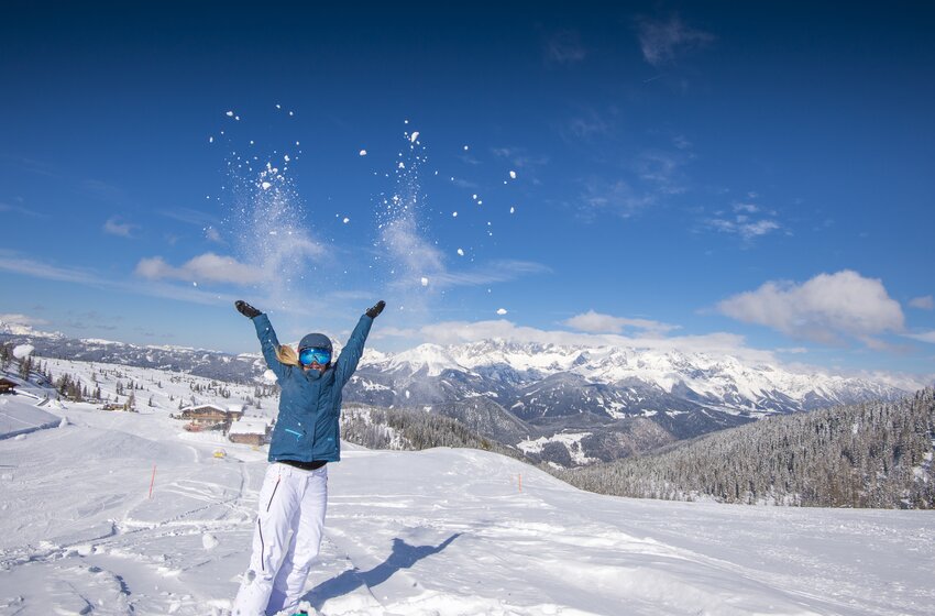 Frau wirft Schnee in die Luft vor verschneiten Bergen und strahlend blauem Himmel. | © Reiteralm Bergbahnen GmbH & Co.KG