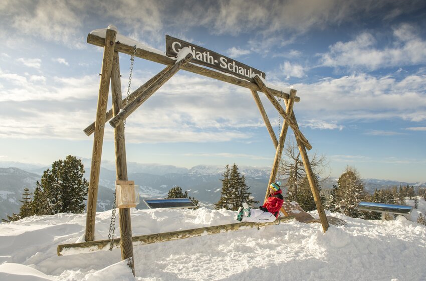 Kind schaukelt auf riesiger Holzschaukel im Schnee mit Blick auf verschneite Berglandschaft. | © Reiteralm Bergbahnen GmbH & Co.KG