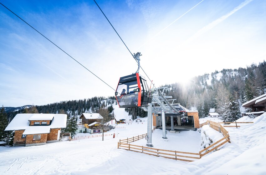Rote Galsterberg-Gondel nahe der Talstation bei Sonnenschein, umgeben von verschneiten Hütten und bewaldeten Hängen. | © Galsterbergalm Bahnen GmbH & Co KG 