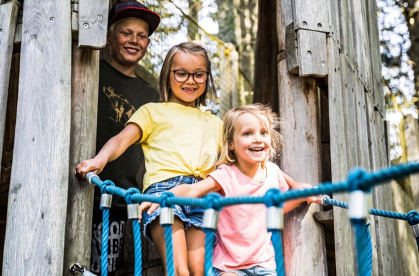 Zwei Mädchen laufen fröhlich über eine Seilbrücke im Wald, ein Jugendlicher steht lächelnd hinter ihnen an einem Holzturm. | © Snow Space Salzburg Bergbahnen