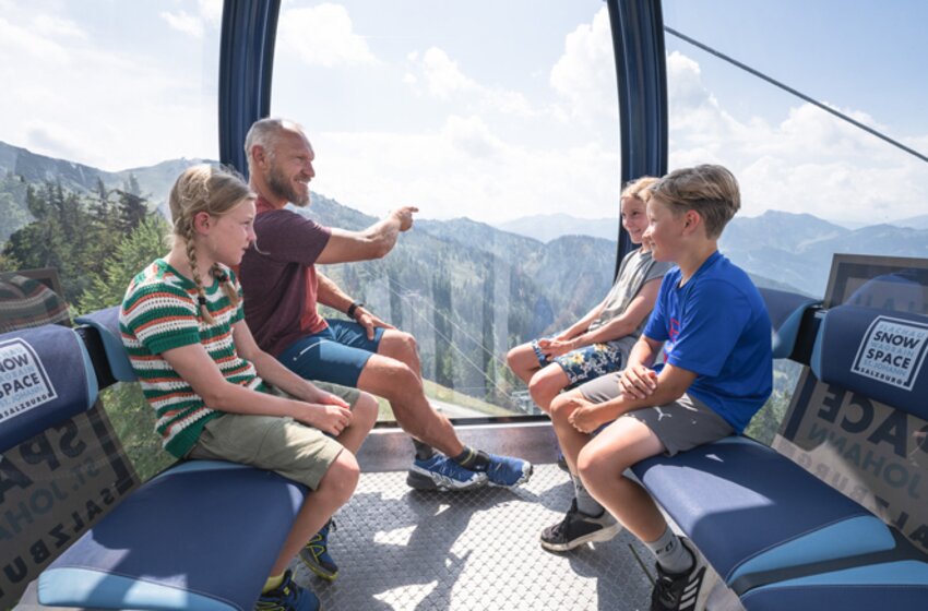 Ein Erwachsener sitzt mit drei Kindern in der Flying Mozart Gondel und blickt auf die sommerliche Berglandschaft draußen. | © Snow Space Salzburg Bergbahnen