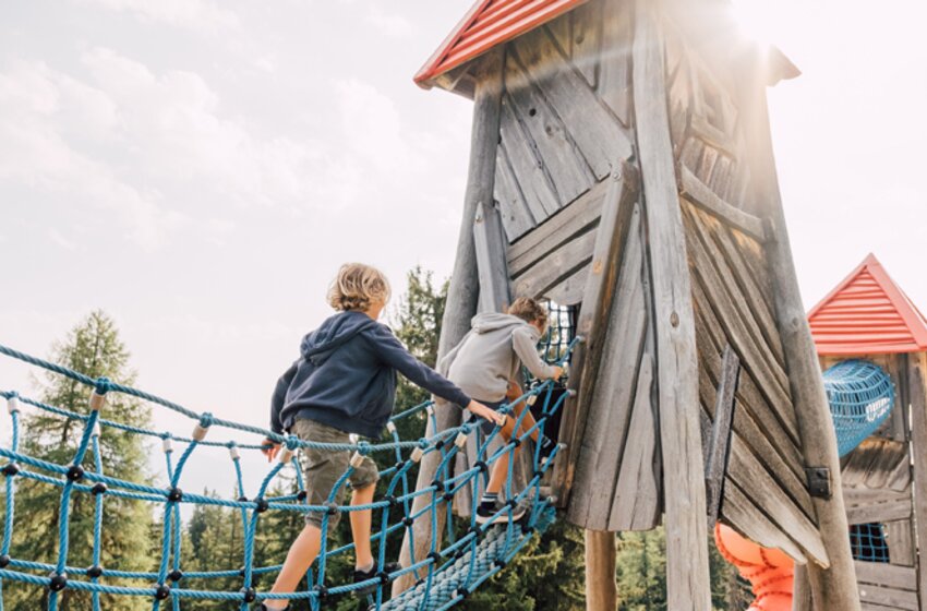 Zwei Kinder klettern über eine blaue Hängebrücke in einen hölzernen Spielturm mit rotem Dach am Geisterberg bei Sonnenschein. | © Snow Space Salzburg Bergbahnen