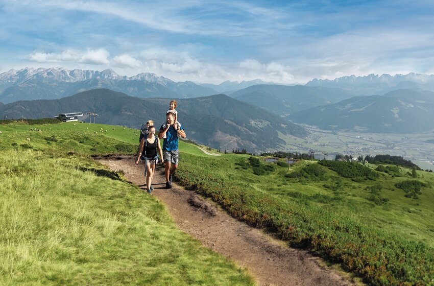 Familie mit zwei Kindern wandert auf einem Bergweg durch grüne Almwiesen vor weitem Alpenpanorama unter blauem Himmel. | © Snow Space Salzburg