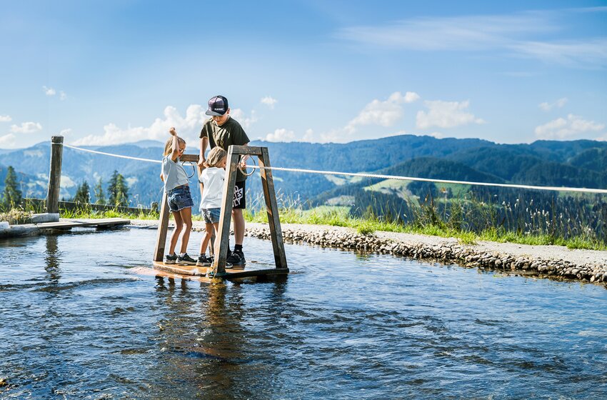Drei Kinder stehen auf einem Wasserfloß in einem flachen Becken vor grünen Hügeln und blauem Sommerhimmel. | © Snow Space Salzburg